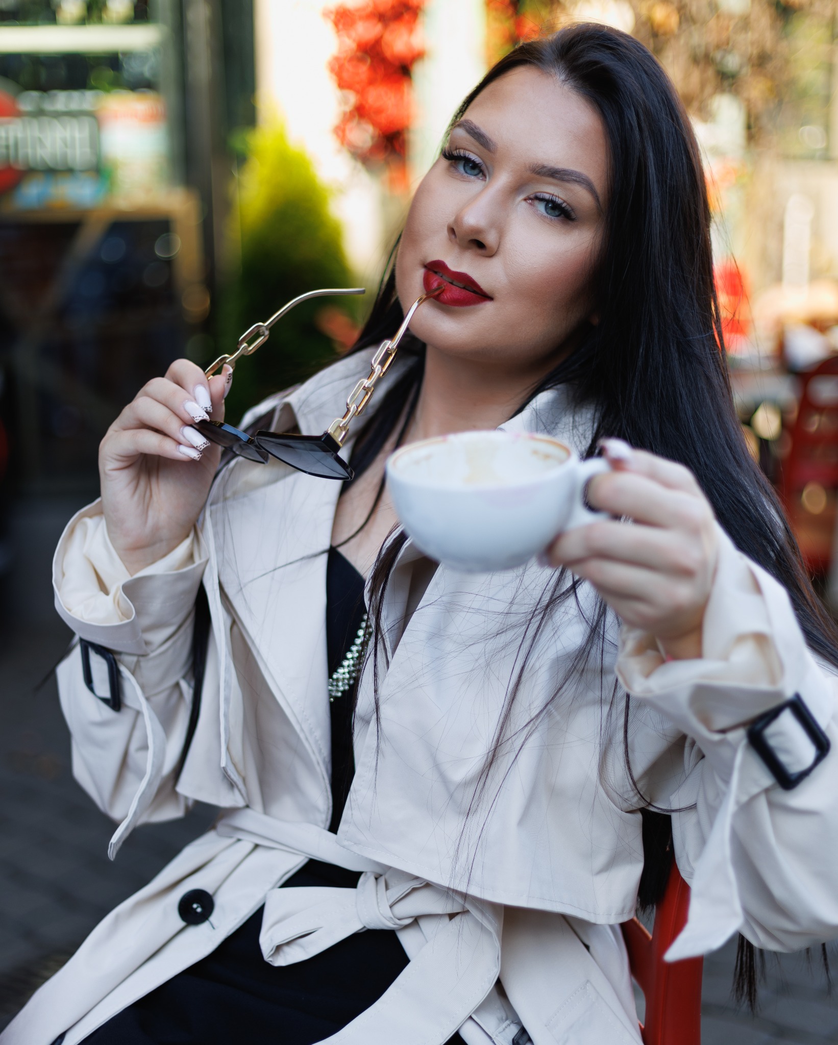 Portret kobiety w beżowym płaszczu, trzymającej filiżankę kawy i okulary przeciwsłoneczne z łańcuszkiem, z czerwonymi ustami i intensywnym makijażem oczu.