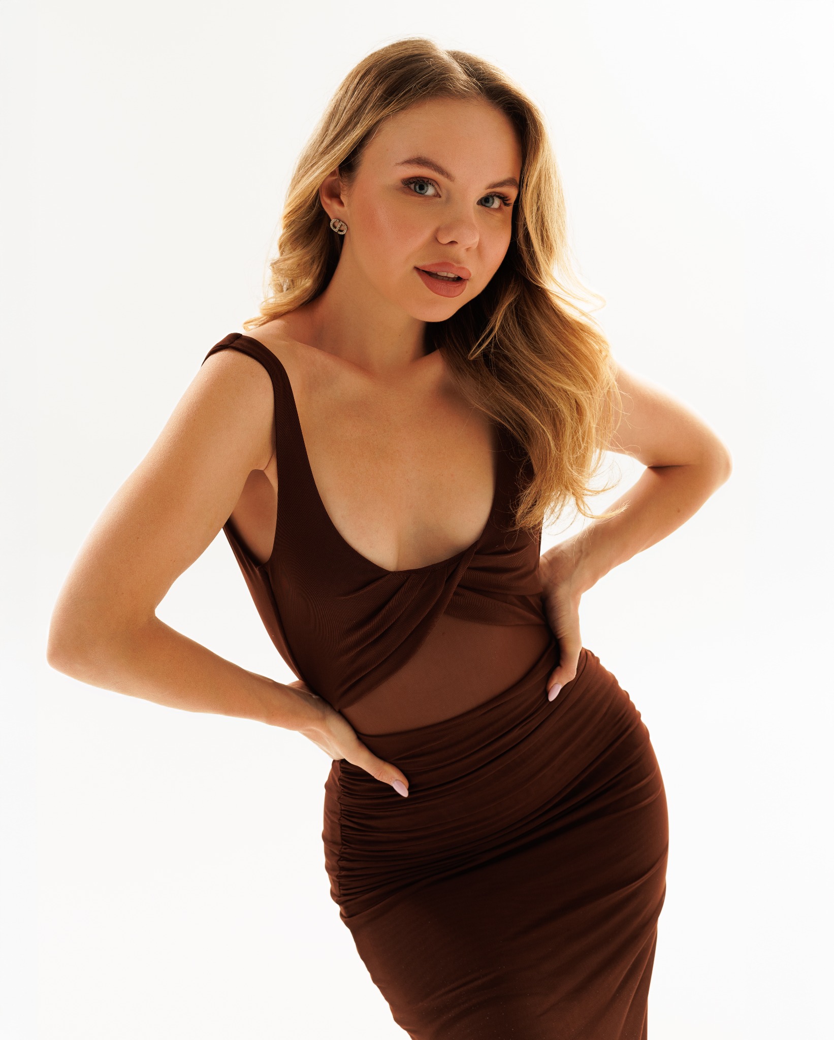 Portret kobiety w brązowej sukience z prześwitującą siateczką, pozującej na białym tle, z delikatnym makijażem i kolczykami