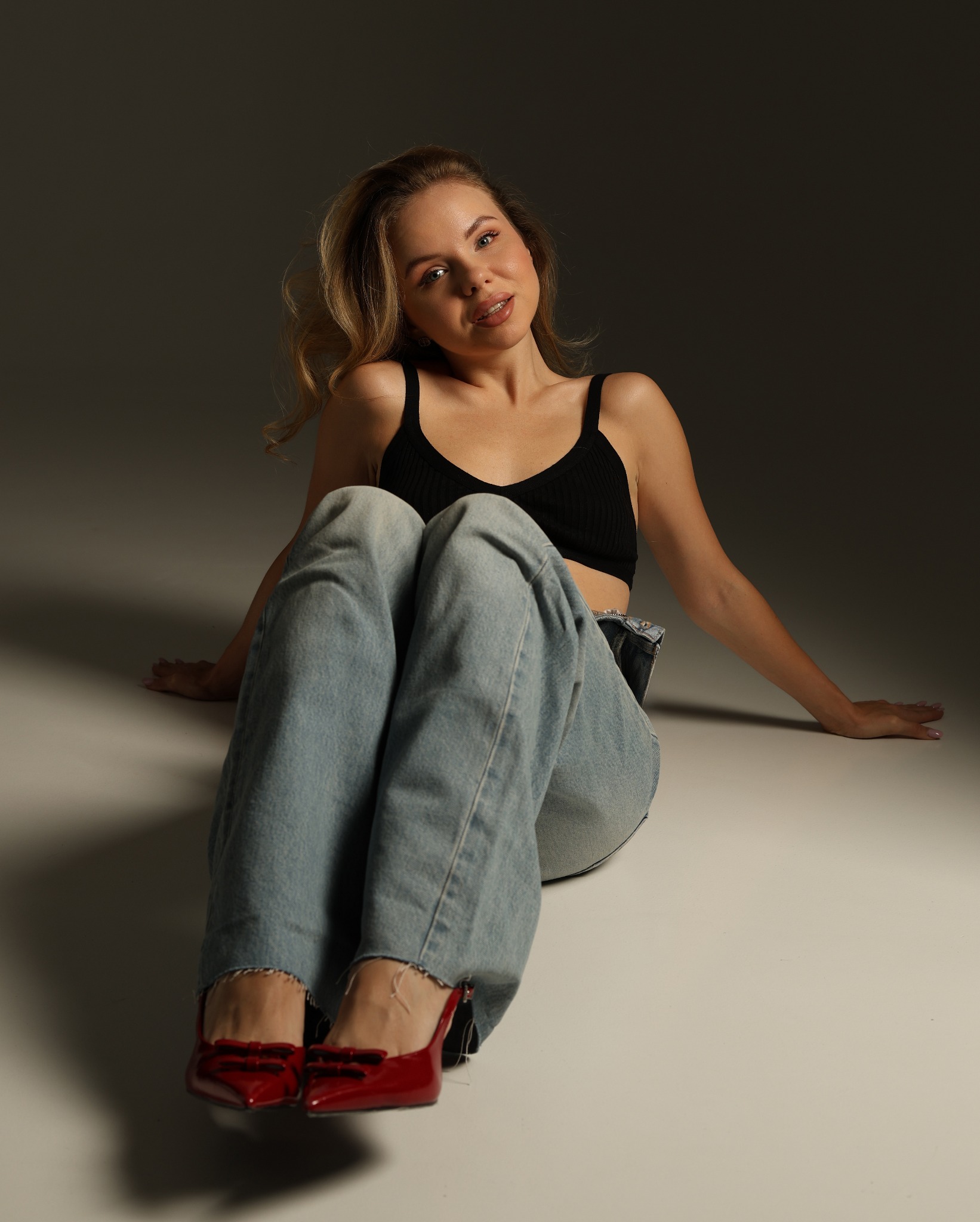 Portret kobiety siedzącej na podłodze w dżinsach i czarnym topie, z czerwonymi lakierowanymi szpilkami z kokardkami, oświetlonej od góry.