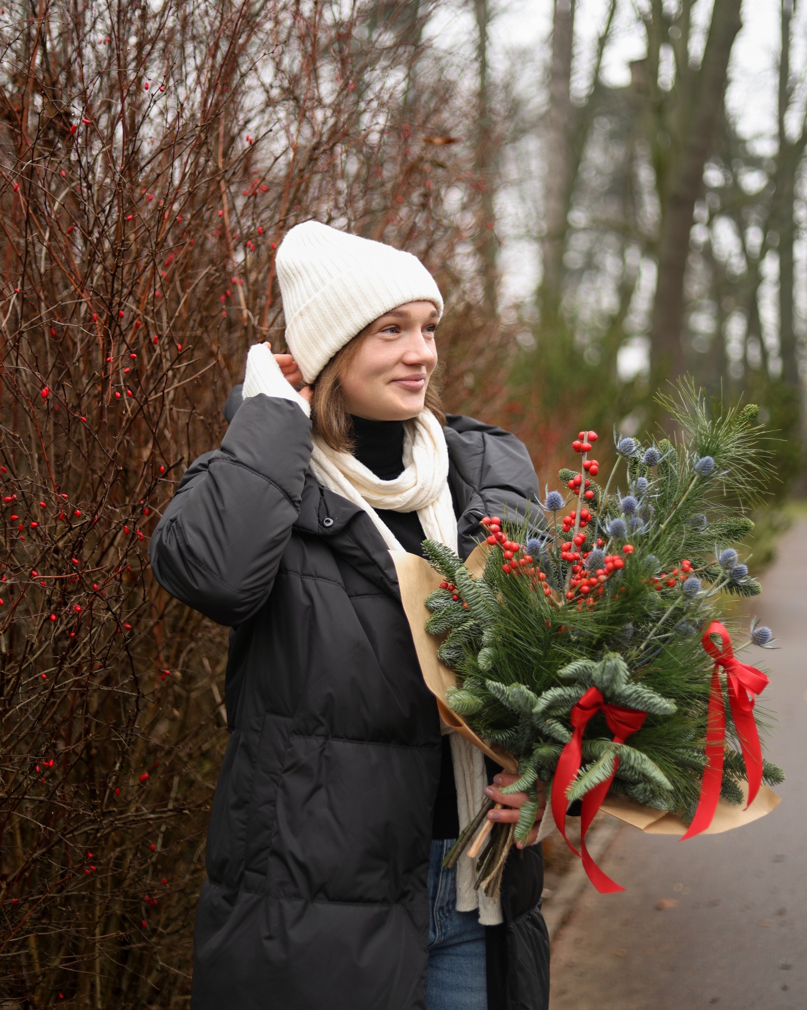 Uśmiechnięta młoda kobieta w zimowej czapce i szaliku trzyma bukiet z gałązek świerku, czerwonych jagód i ostu na tle krzewu z czerwonymi owocami.