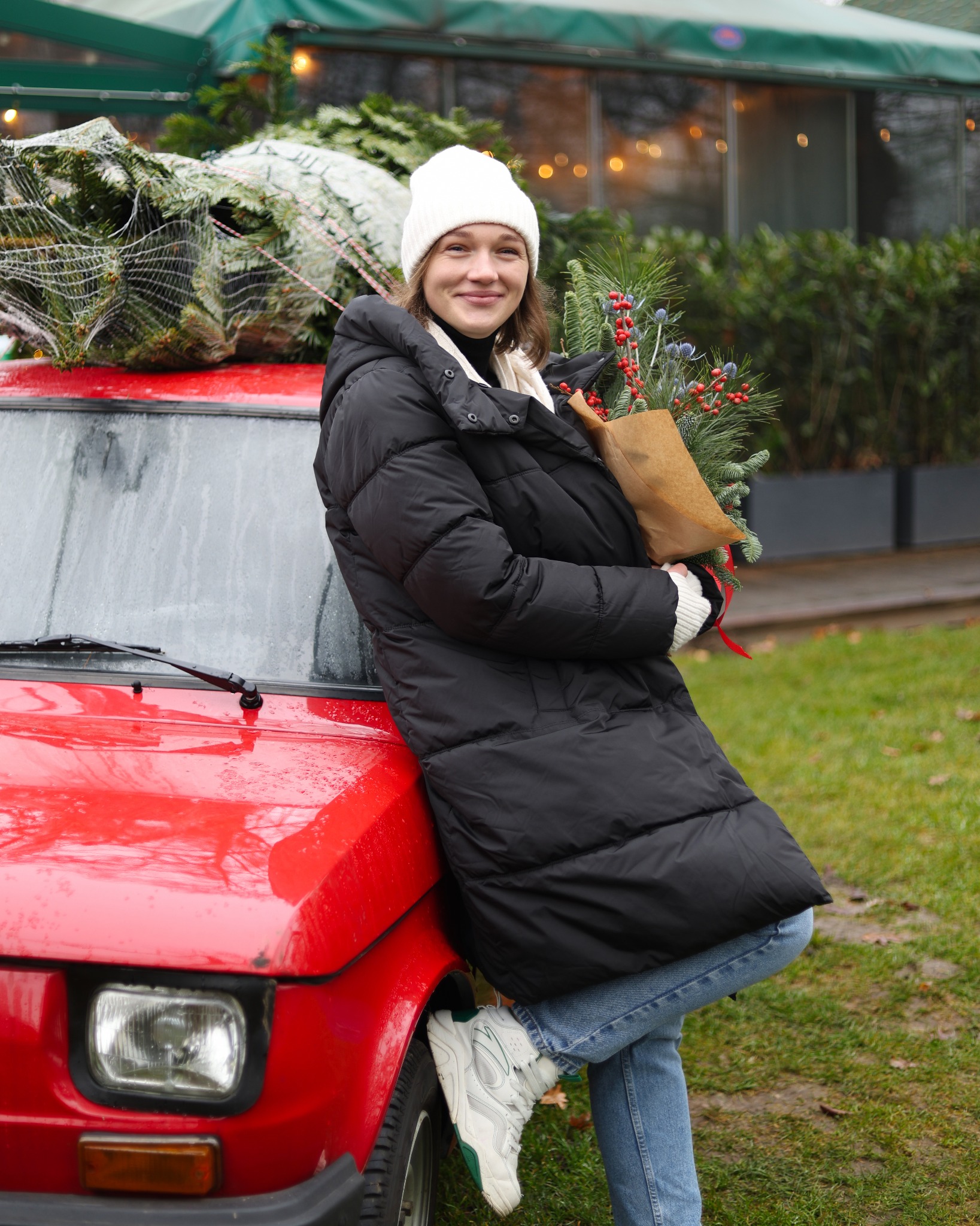 Uśmiechnięta kobieta w zimowej kurtce i czapce, oparta o czerwony samochód, trzyma bukiet z gałązek iglastych i czerwonych jagód, na dachu auta choinka w siatce.