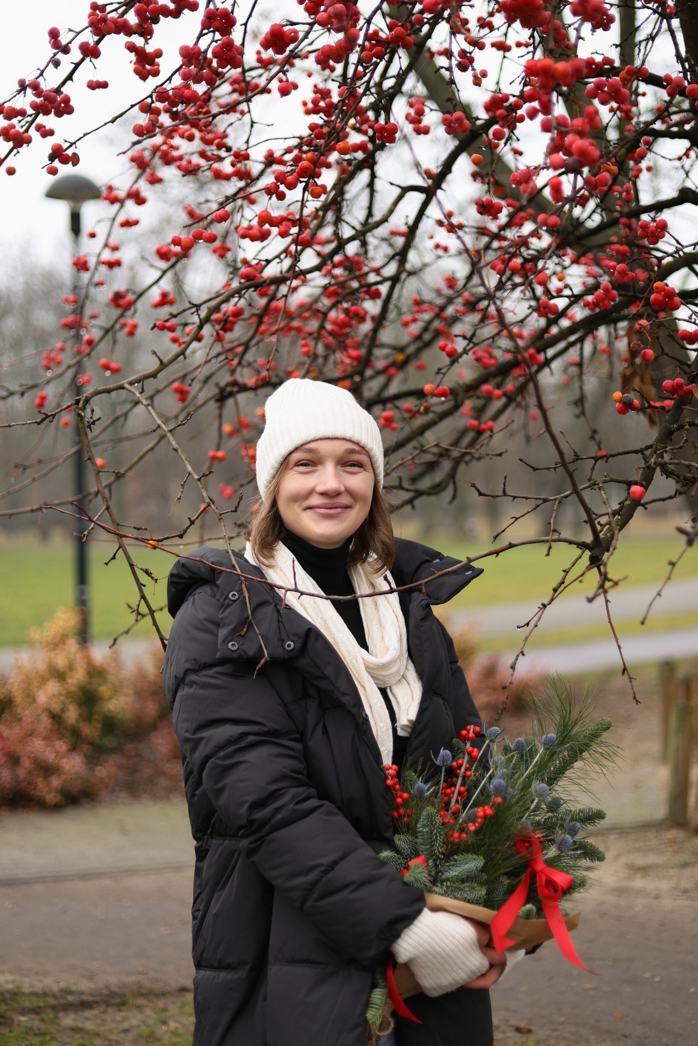 Portret uśmiechniętej kobiety w zimowej stylizacji z bukietem z gałązek i czerwonych jagód na tle drzewa z czerwonymi owocami w Grodzisku Mazowieckim.