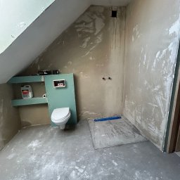 DAW-MAT Dawid Hanuszek - Łazienka w trakcie remontu w poddaszu: surowe ściany z resztkami gładzi, zamontowana toaleta na tle zielonej płyty, widoczne rury instalacji, grunt na półce i betonowa wylewka na podłodze.