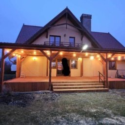 DAW-MAT Dawid Hanuszek - Zadaszony taras drewniany z oświetleniem żarówkowym przy domu jednorodzinnym, widok z ogrodu, wieczorne oświetlenie.
