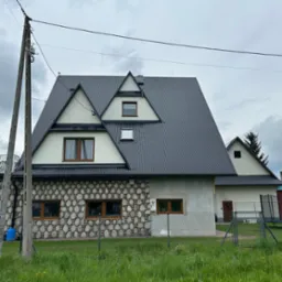 Dom z ciemnym, blachodachówkowym dachem i elewacją z kamienną okładziną. Widoczne okna z drewnianą stolarką i zielony trawnik przed ogrodzeniem.
