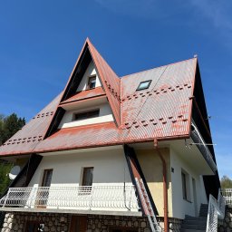 DAW-MAT Dawid Hanuszek - Dom z dachem z blachy trapezowej, częściowo pomalowanym na czerwono. Widoczna drabina oparta o ścianę, sugerująca trwające prace. Jasne niebo w tle.