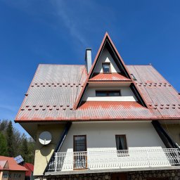 DAW-MAT Dawid Hanuszek - Dwuspadowy dach z blachy trapezowej, częściowo pokryty rdzą, z widocznym kominem i oknami poddasza na tle błękitnego nieba. Ujęcie od dołu.