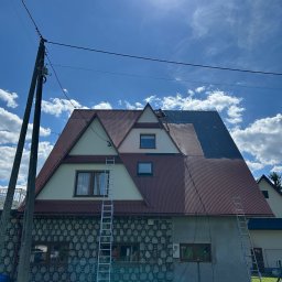 DAW-MAT Dawid Hanuszek - Dom z częściowo pomalowanym dachem w odcieniach brązu i szarości, drabiny opierają się o budynek, widoczna osoba podczas pracy na dachu.