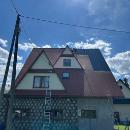 DAW-MAT Dawid Hanuszek - Dwuspadowy dach z blachy trapezowej, częściowo pomalowany na szaro. Na dachu stoi osoba. Dwie drabiny opierają się o budynek. Częściowo kamienna elewacja.