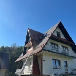 Dwupiętrowy dom z metalowym dachem w trakcie czyszczenia przez pracownika z użyciem myjki ciśnieniowej. Widoczna drabina oparta o ścianę. Błękitne niebo w tle.