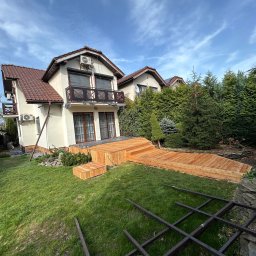 DAW-MAT Dawid Hanuszek - Nowy taras z drewna przy domu z balkonem. Widoczne schody tarasowe i fragment konstrukcji pergoli. Jasne drewno kontrastuje z zielenią trawnika i iglaków.