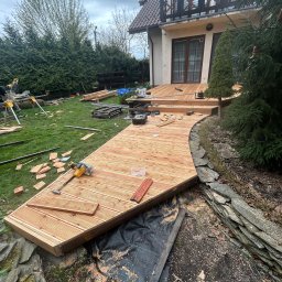 DAW-MAT Dawid Hanuszek - Budowa tarasu drewnianego: zakrzywiona konstrukcja z desek, narzędzia pracy na trawie, dom w tle. Widoczne etapy montażu i przygotowania terenu.