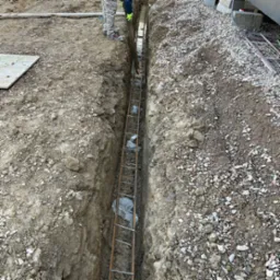 Wykop zbrojony stalą i podkładami z bloczków betonowych. Widoczne fragmenty odzieży robotników i podłoże z ziemi i kamieni. Przygotowanie do wylewania fundamentów.