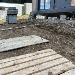 DAW-MAT Dawid Hanuszek - Przygotowanie terenu pod budowę: zbrojenie fundamentów w wykopie, palety i bloczki betonowe, dom modułowy na betonowych podporach w tle.