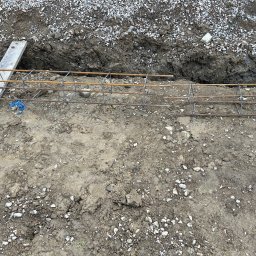 DAW-MAT Dawid Hanuszek - Zbrojenie fundamentu tarasu na gruncie, przygotowane do zalania betonem. Widoczne pręty zbrojeniowe połączone drutem, wykop i podsypka żwirowa.