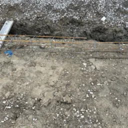 Zbrojenie fundamentu tarasu na gruncie, przygotowane do zalania betonem. Widoczne pręty zbrojeniowe połączone drutem, wykop i podsypka żwirowa.