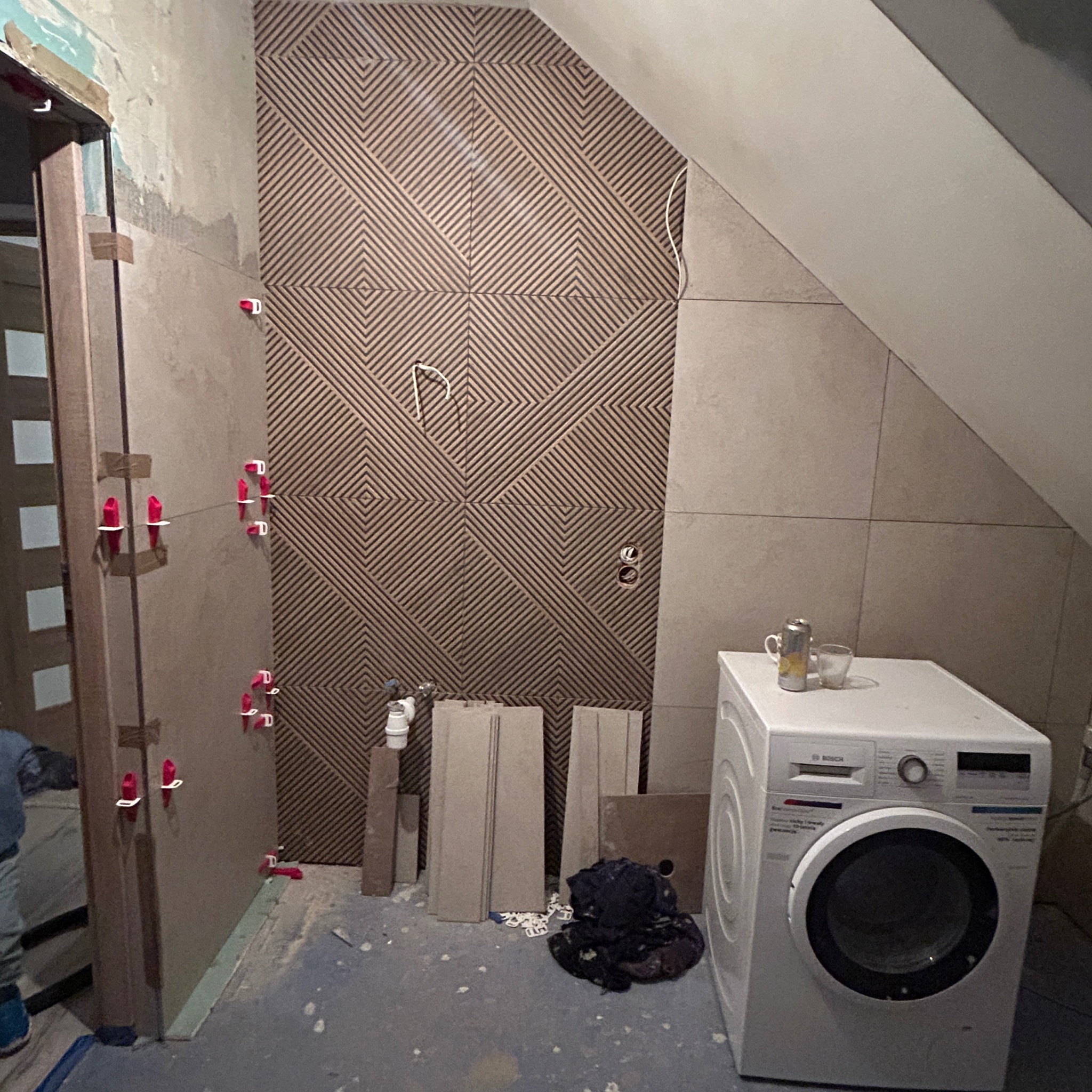 Montaż płytek z geometrycznym wzorem na ścianie w łazience na poddaszu, widoczne narzędzia glazurnicze, pralka i niedokończone prace wykończeniowe.