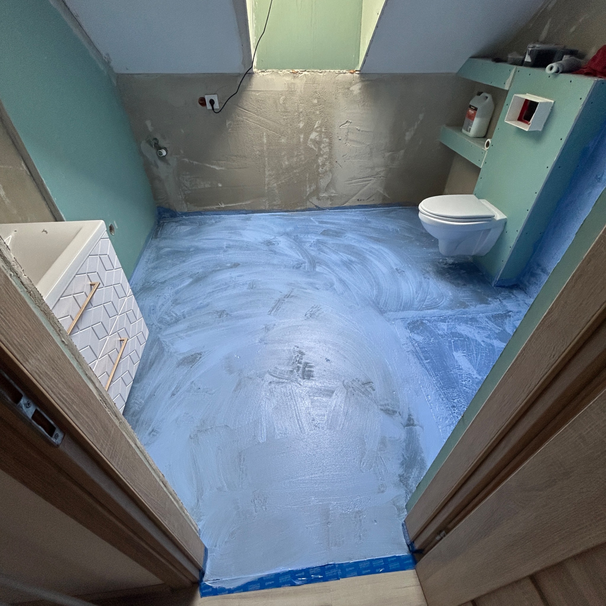 Łazienka na poddaszu w trakcie remontu, z położoną wylewką samopoziomującą na podłodze, częściowo obłożona płytami kartonowo-gipsowymi, widoczna umywalka z mozaiką i toaleta pod skosem dachu.