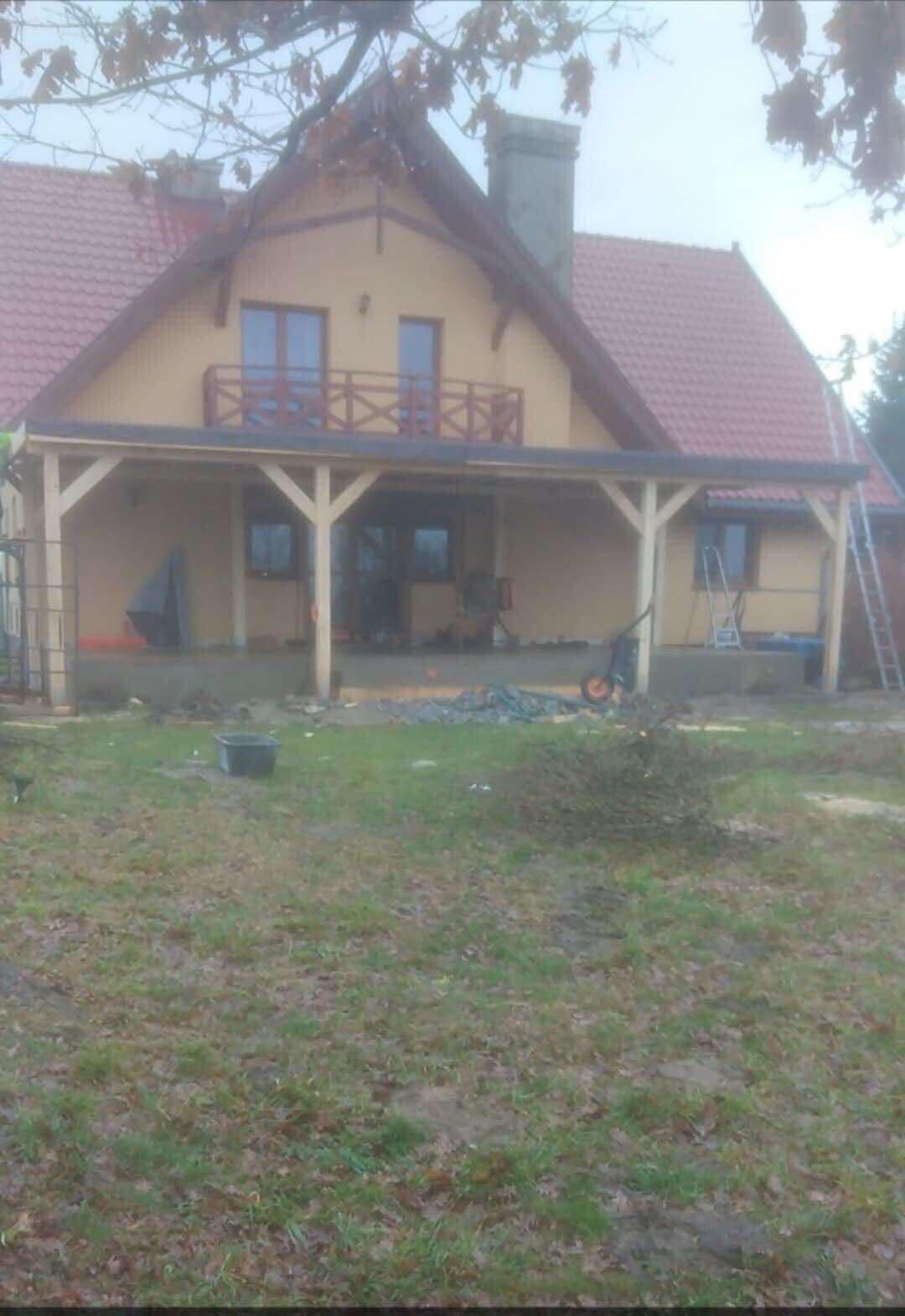 Dom jednorodzinny z zadaszonym tarasem w trakcie budowy, widoczne materiały budowlane i narzędzia, trawnik z mokrą trawą.