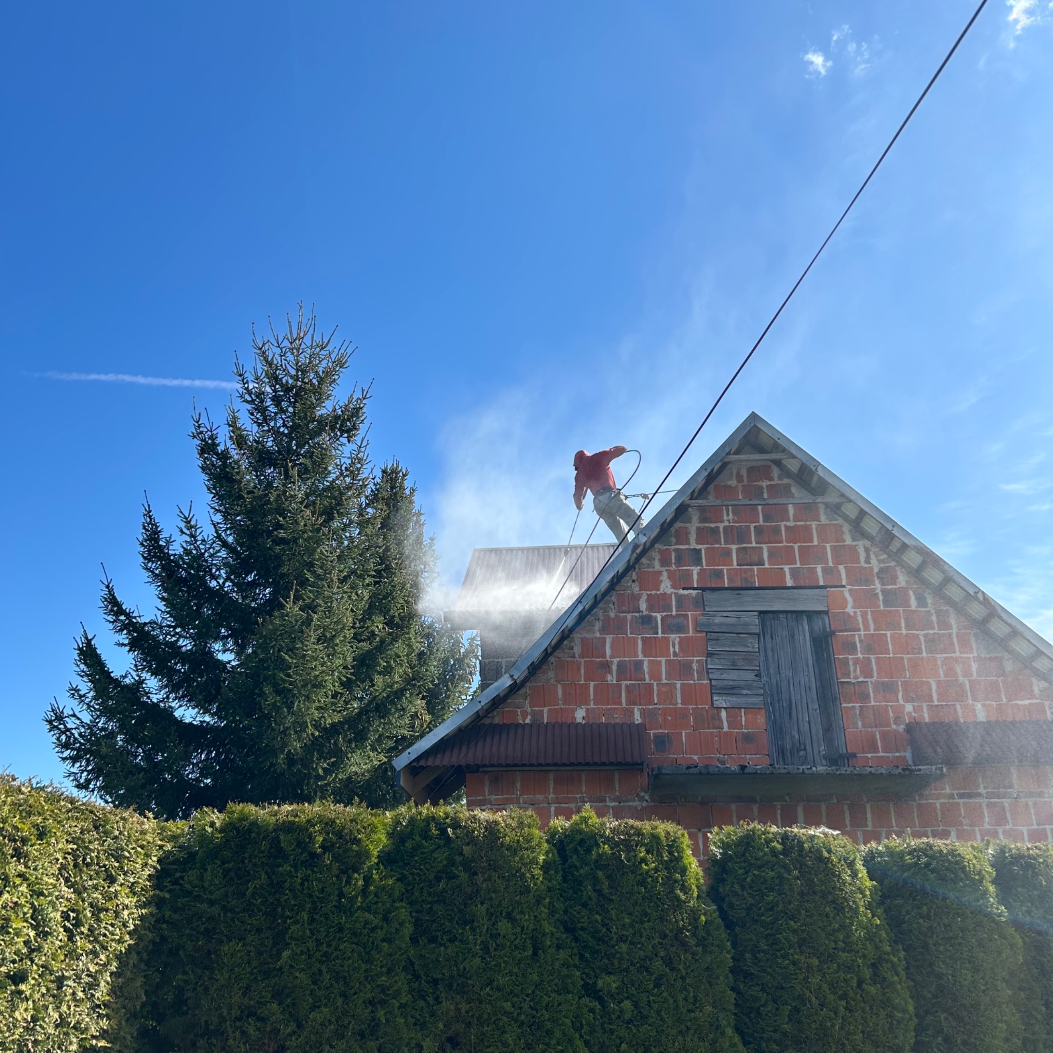 Mężczyzna w czerwonej bluzie myje dach ceglanego domu pod silnym strumieniem wody z węża, na tle błękitnego nieba i zielonych drzew.
