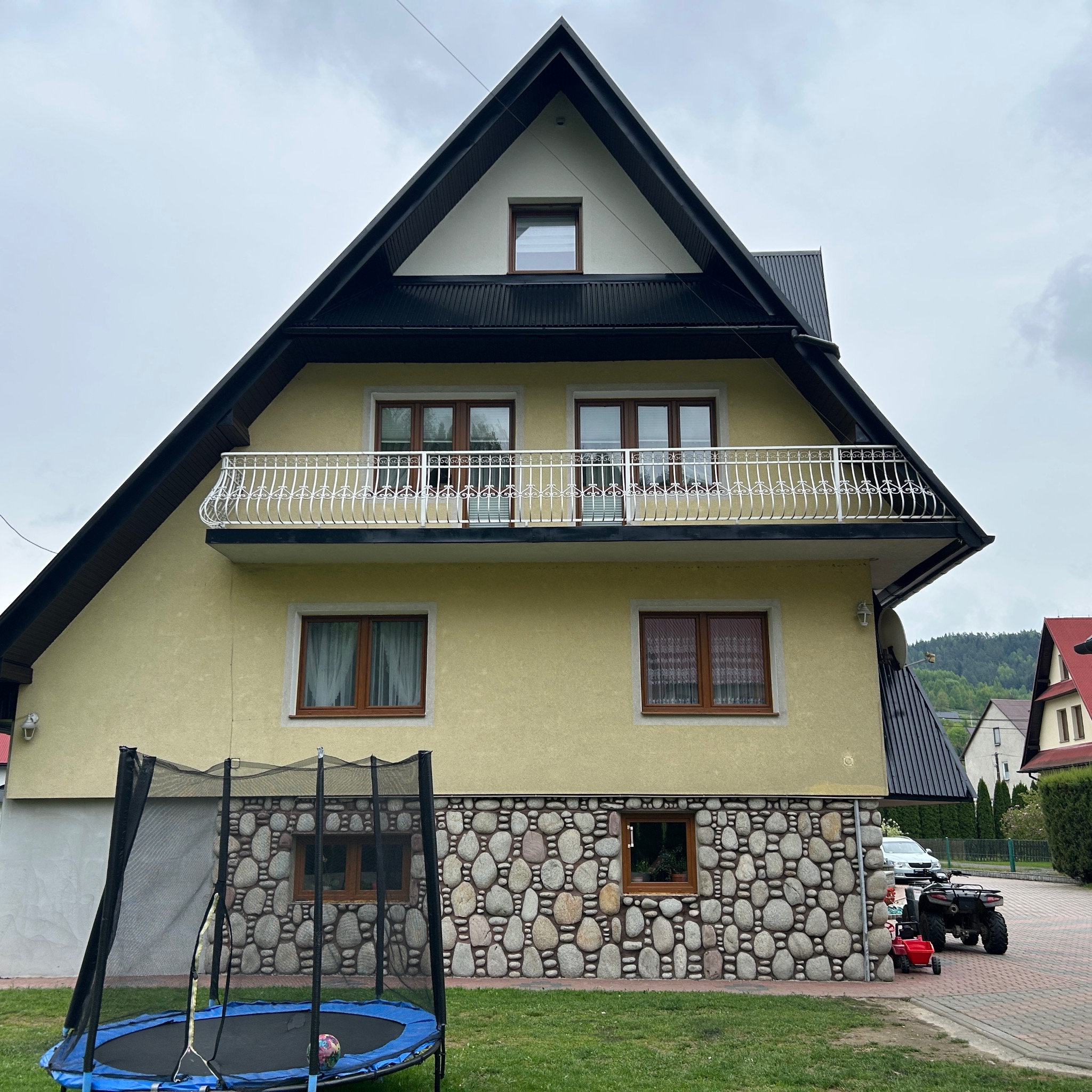 Dwupiętrowy dom z żółtą elewacją, czarnym dachem i kamiennym wykończeniem parteru. Biała balustrada balkonu. Na trawniku trampolina.