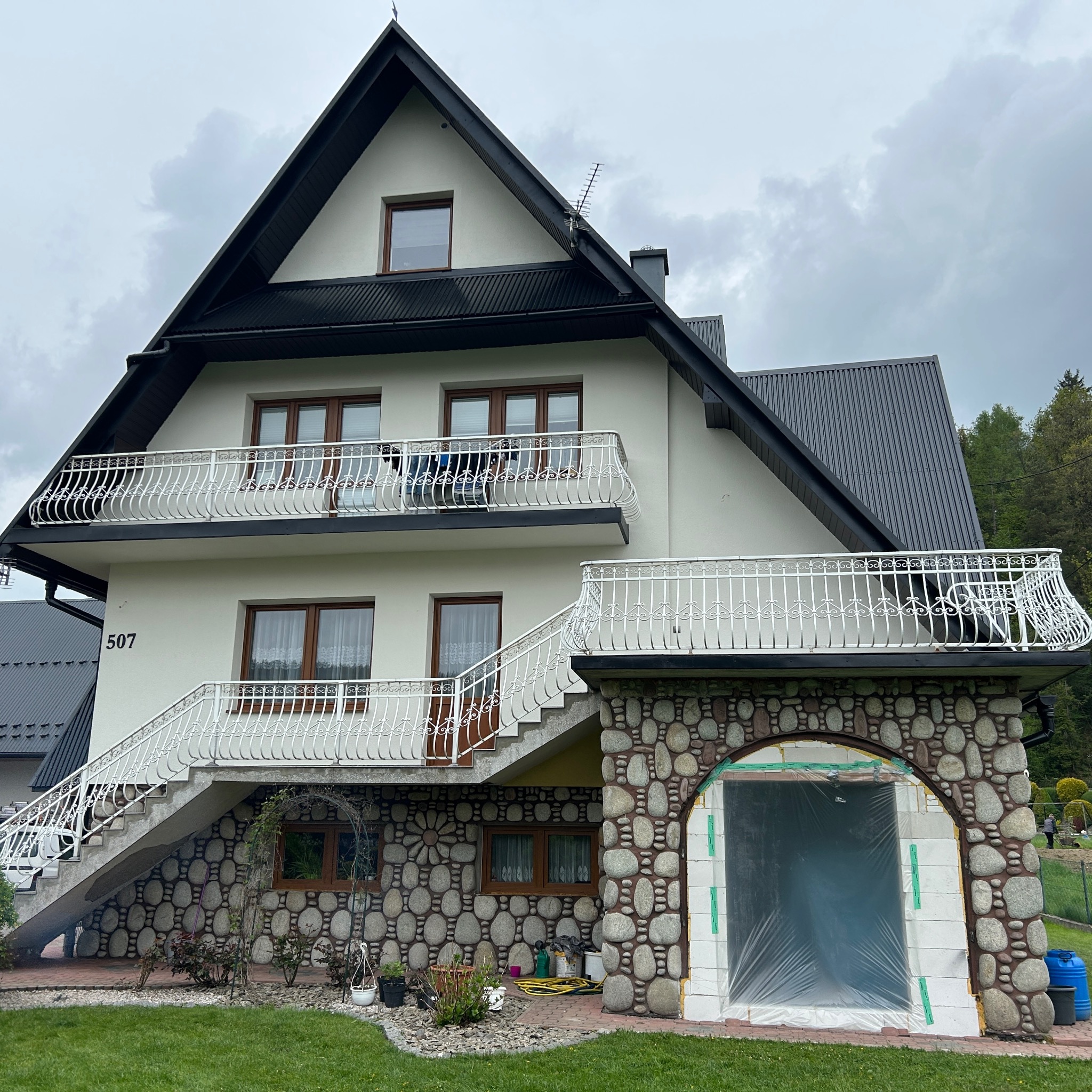 Dwupiętrowy dom z ciemnym dachem i białymi balkonami. Kamienna elewacja z łukowym wejściem zabezpieczonym folią. Numer domu 507 widoczny na ścianie.