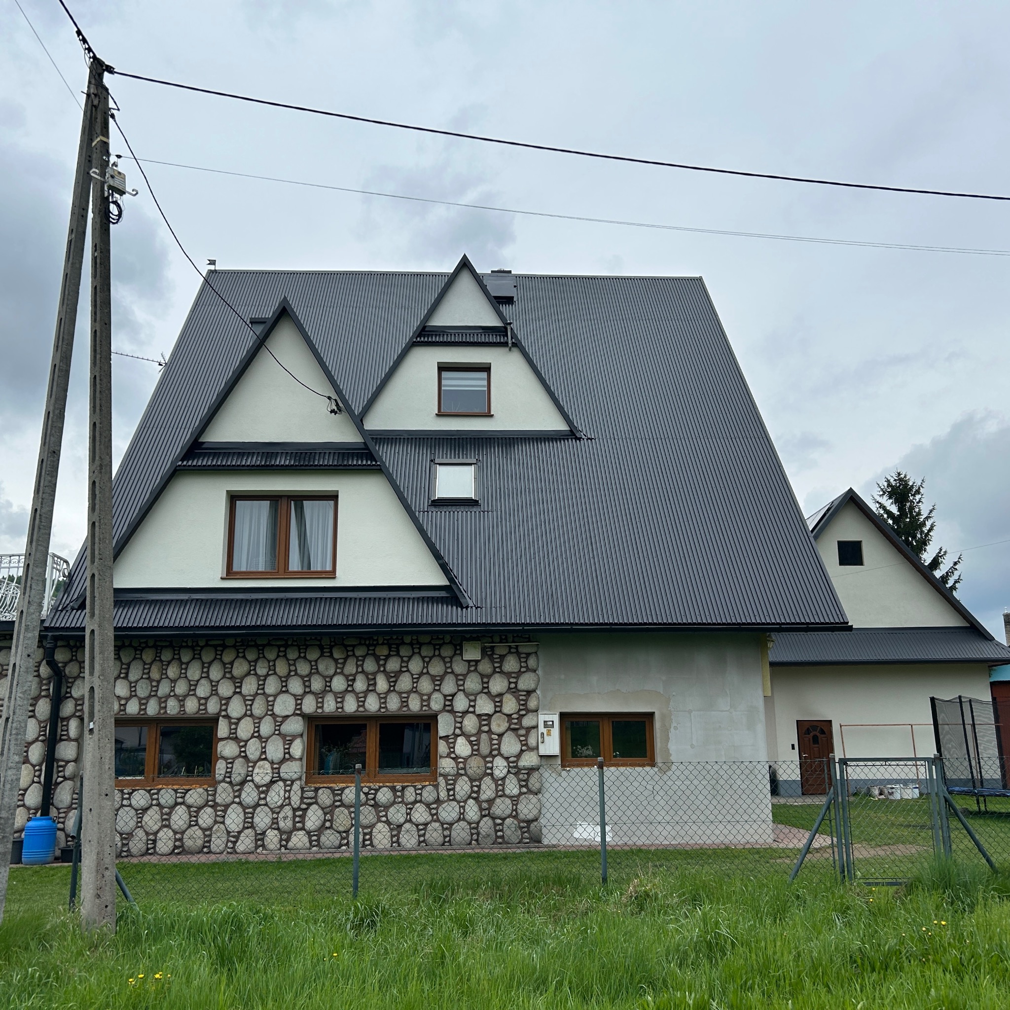Dom z ciemnym, blachodachówkowym dachem i elewacją z kamienną okładziną. Widoczne okna z drewnianą stolarką i zielony trawnik przed ogrodzeniem.