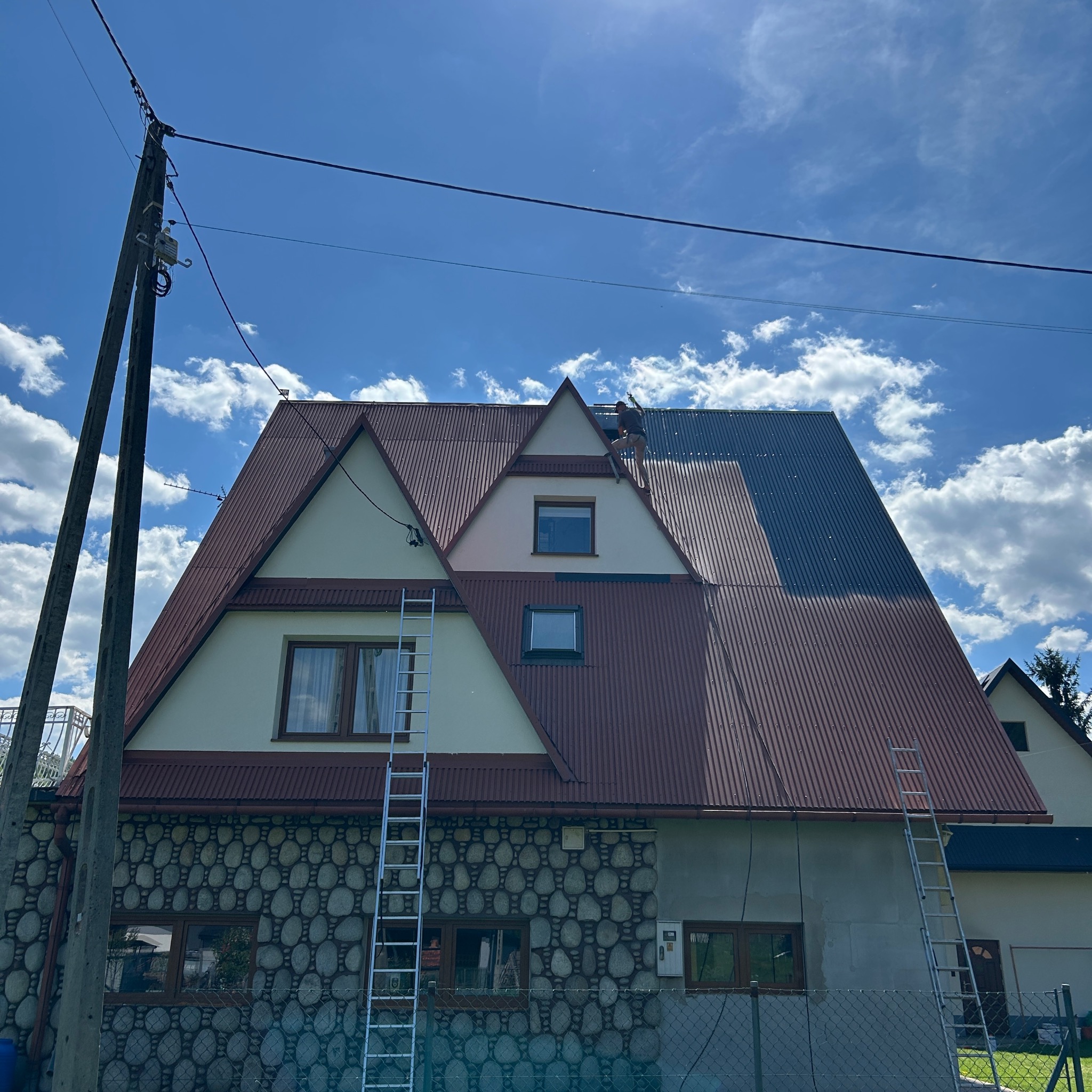 Dwuspadowy dach z blachy trapezowej, częściowo pomalowany na szaro. Na dachu stoi osoba. Dwie drabiny opierają się o budynek. Częściowo kamienna elewacja.