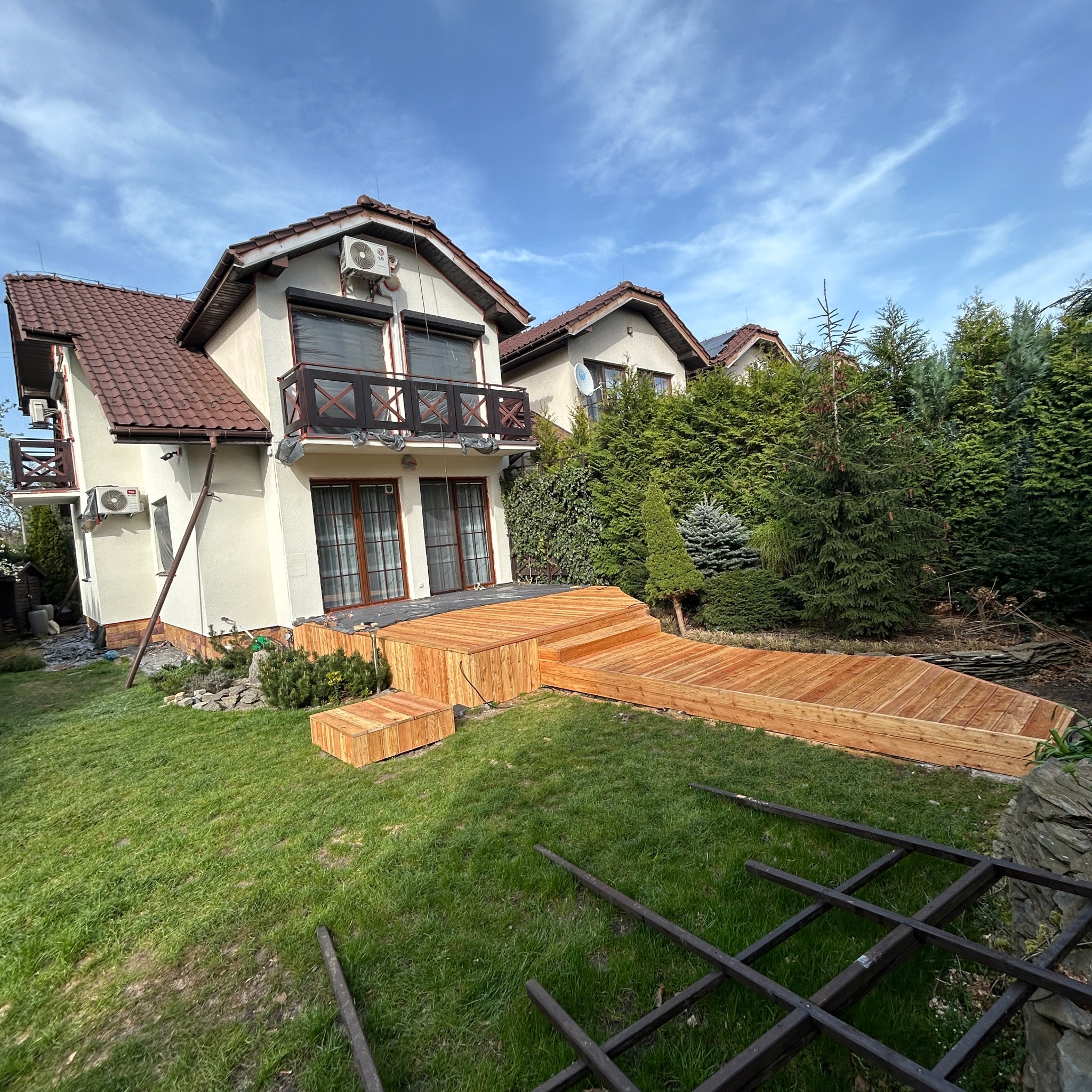 Nowy taras z drewna przy domu z balkonem. Widoczne schody tarasowe i fragment konstrukcji pergoli. Jasne drewno kontrastuje z zielenią trawnika i iglaków.