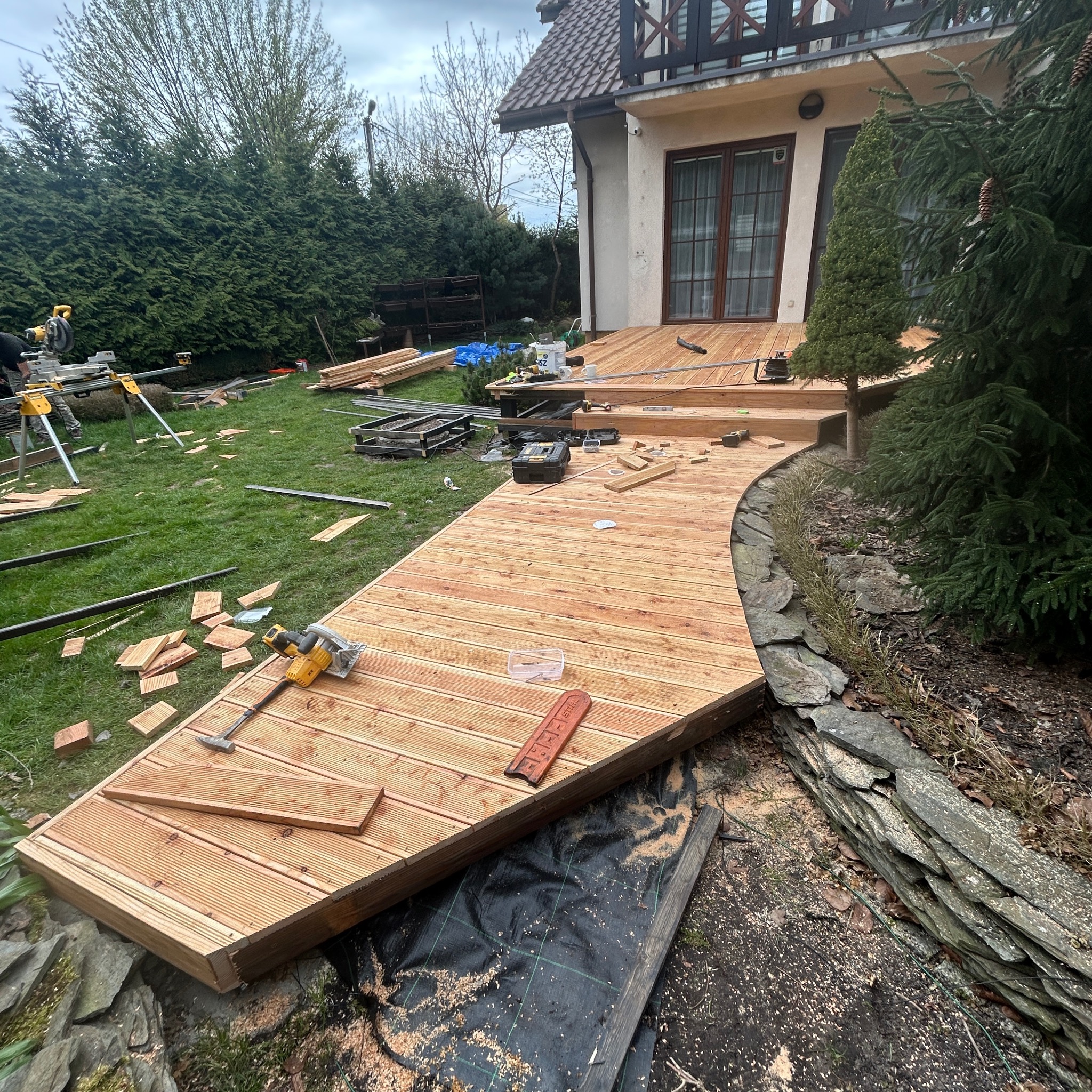 Budowa tarasu drewnianego: zakrzywiona konstrukcja z desek, narzędzia pracy na trawie, dom w tle. Widoczne etapy montażu i przygotowania terenu.