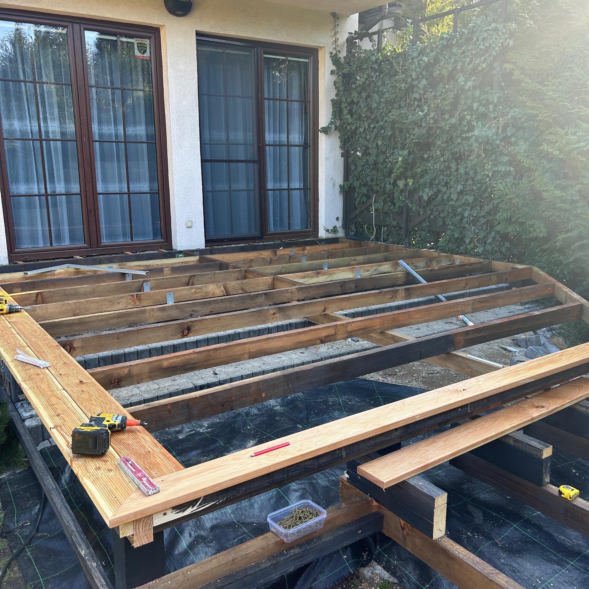 Budowa tarasu drewnianego: drewniana konstrukcja szkieletowa z częściowo zamontowanymi deskami, wkrętarka i narzędzia na budowie przy domu z oknami.