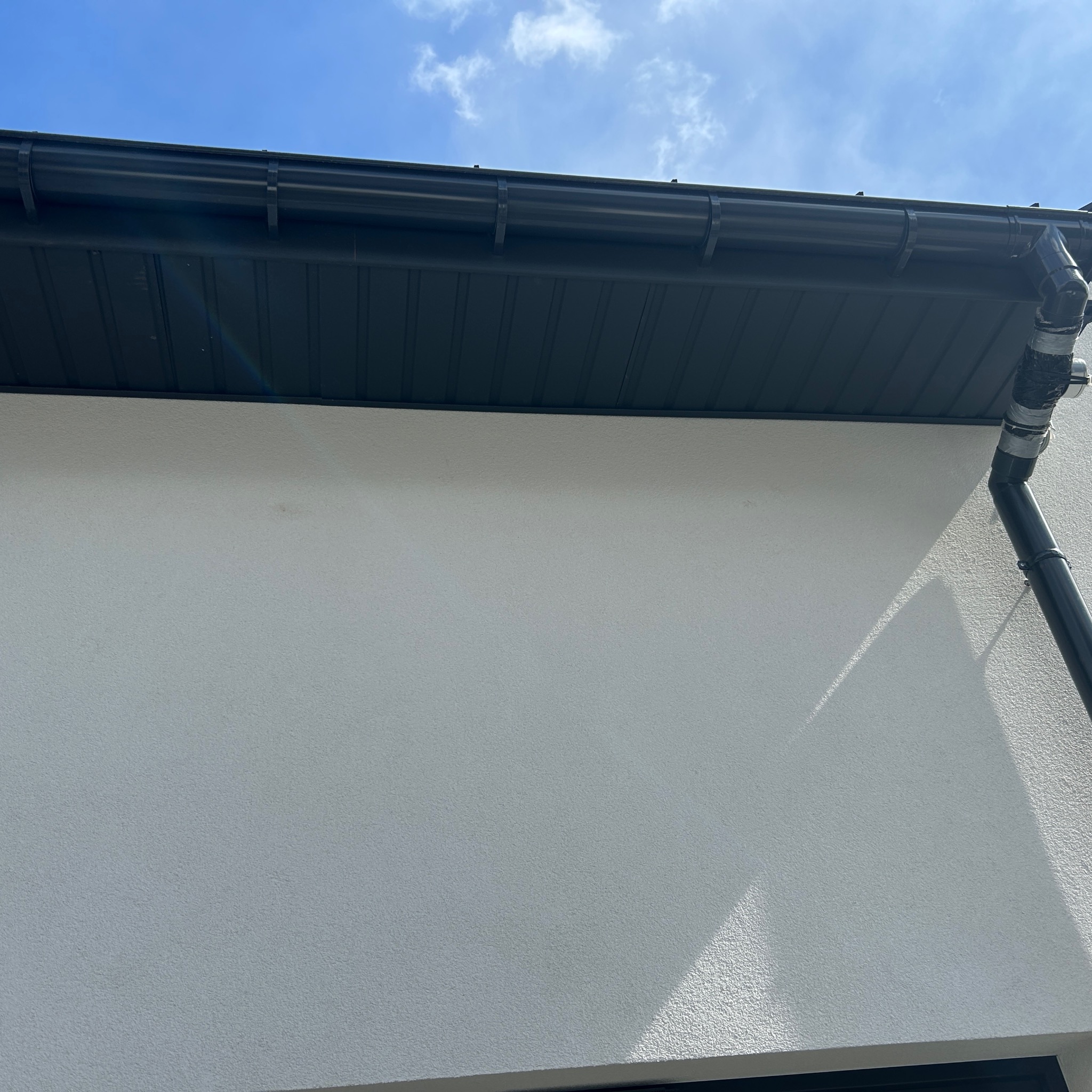 Fragment elewacji z zamontowaną, czarną rynną i rurą spustową, widoczne mocowania. Ściana o jasnej, chropowatej fakturze w słoneczny dzień.