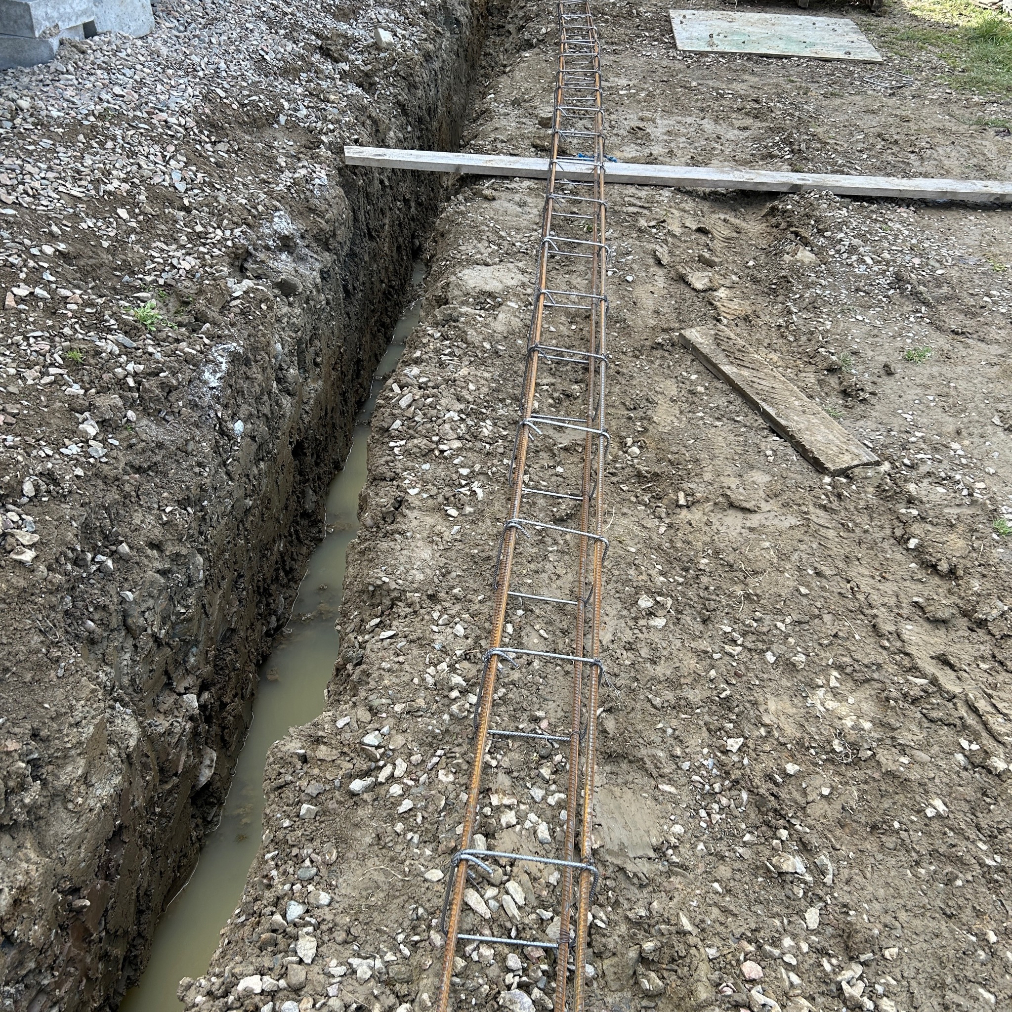 Ziemny wykop z wodą i zbrojeniem, przygotowany pod fundament. Widoczne elementy konstrukcyjne i grunt. Ujęcie z góry ukazuje detale wykopu i zbrojenia.