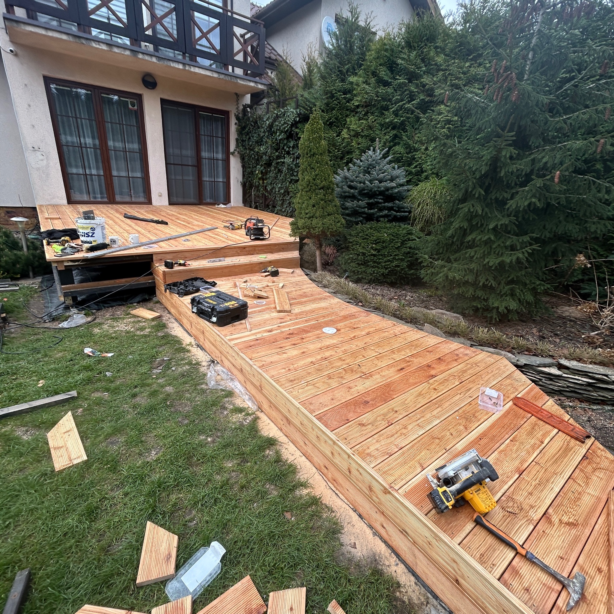 Budowa tarasu z drewna: deski ułożone, narzędzia pracy widoczne. Częściowo ukończony taras przy domu z ogrodem w tle. Widoczne resztki materiałów i narzędzia.