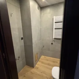 Nowoczesna łazienka z szarymi, kwadratowymi płytkami na ścianach, drewnopodobną podłogą i białym grzejnikiem drabinkowym, widziana zza ciemnych drzwi.