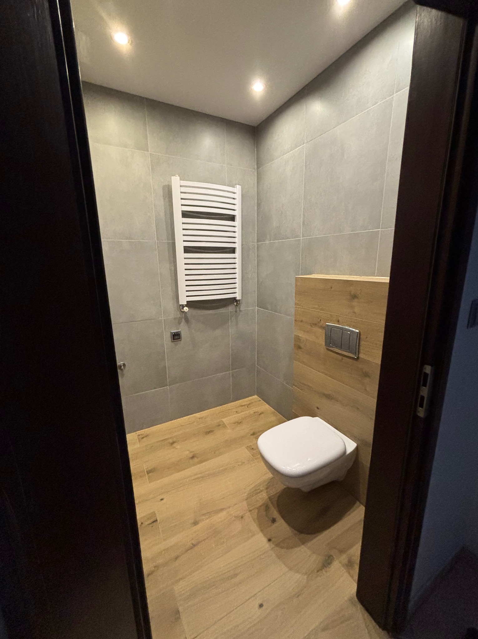 Nowoczesna łazienka z szarymi płytkami, drewnopodobną podłogą, podwieszaną toaletą i białym grzejnikiem drabinkowym, widok zza ciemnych drzwi.