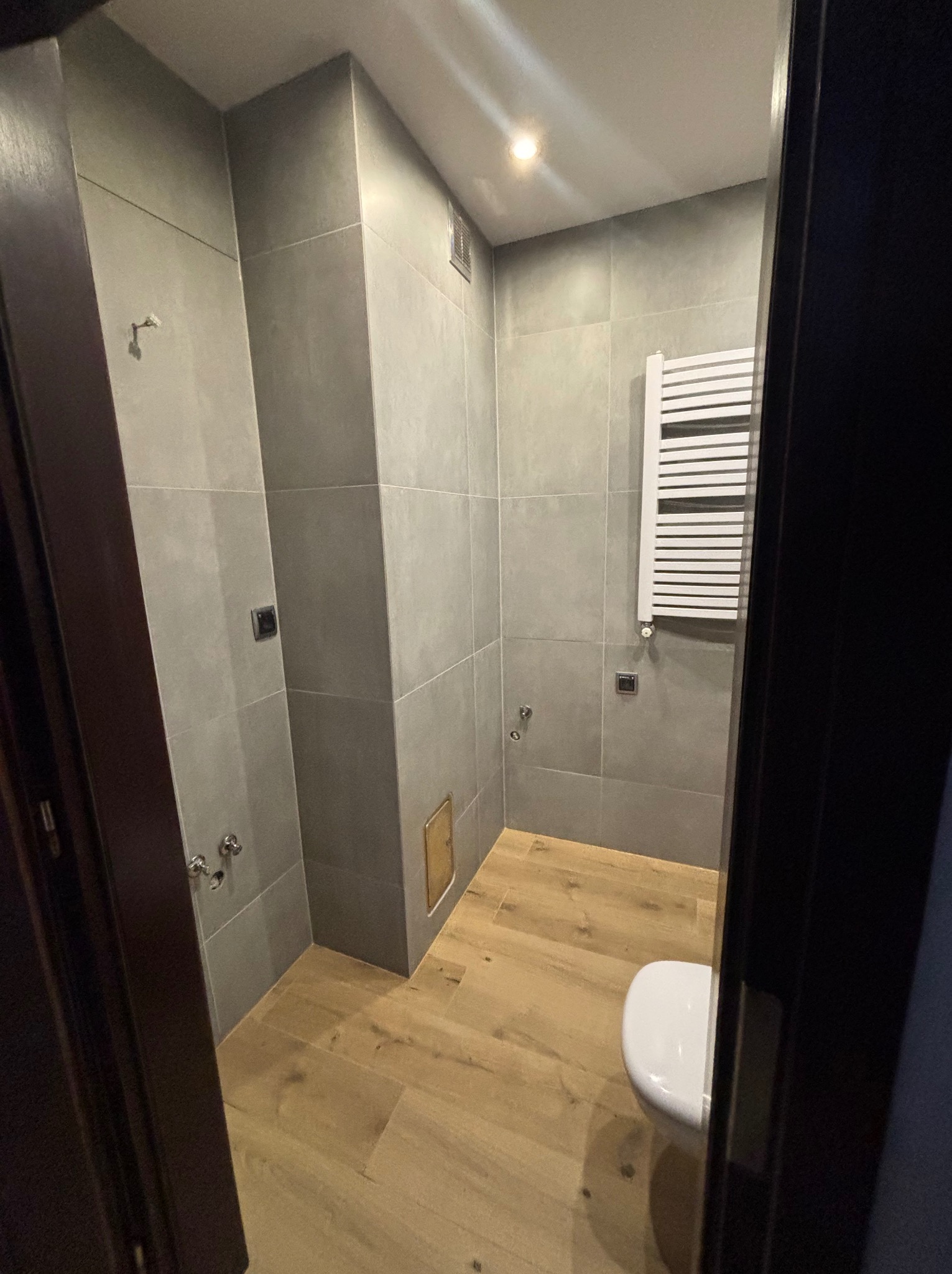 Nowoczesna łazienka z szarymi, kwadratowymi płytkami na ścianach, drewnopodobną podłogą i białym grzejnikiem drabinkowym, widziana zza ciemnych drzwi.