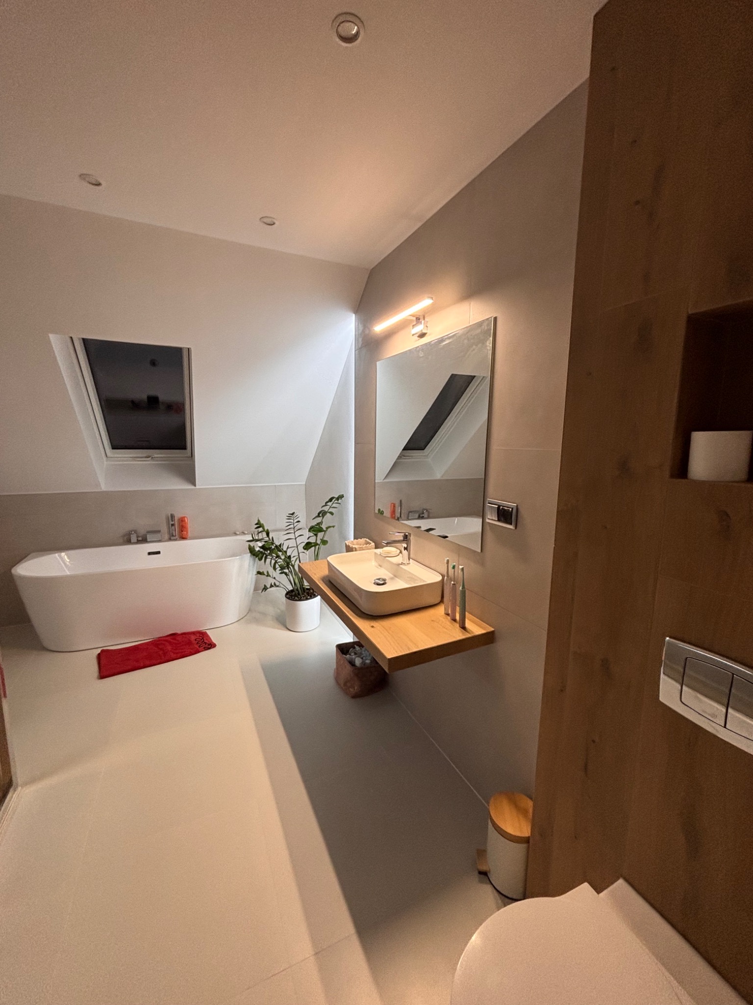 Jasna łazienka na poddaszu z wanną, umywalką na drewnianym blacie i oknem dachowym. Minimalistyczny wystrój z rośliną doniczkową i akcentami drewna.