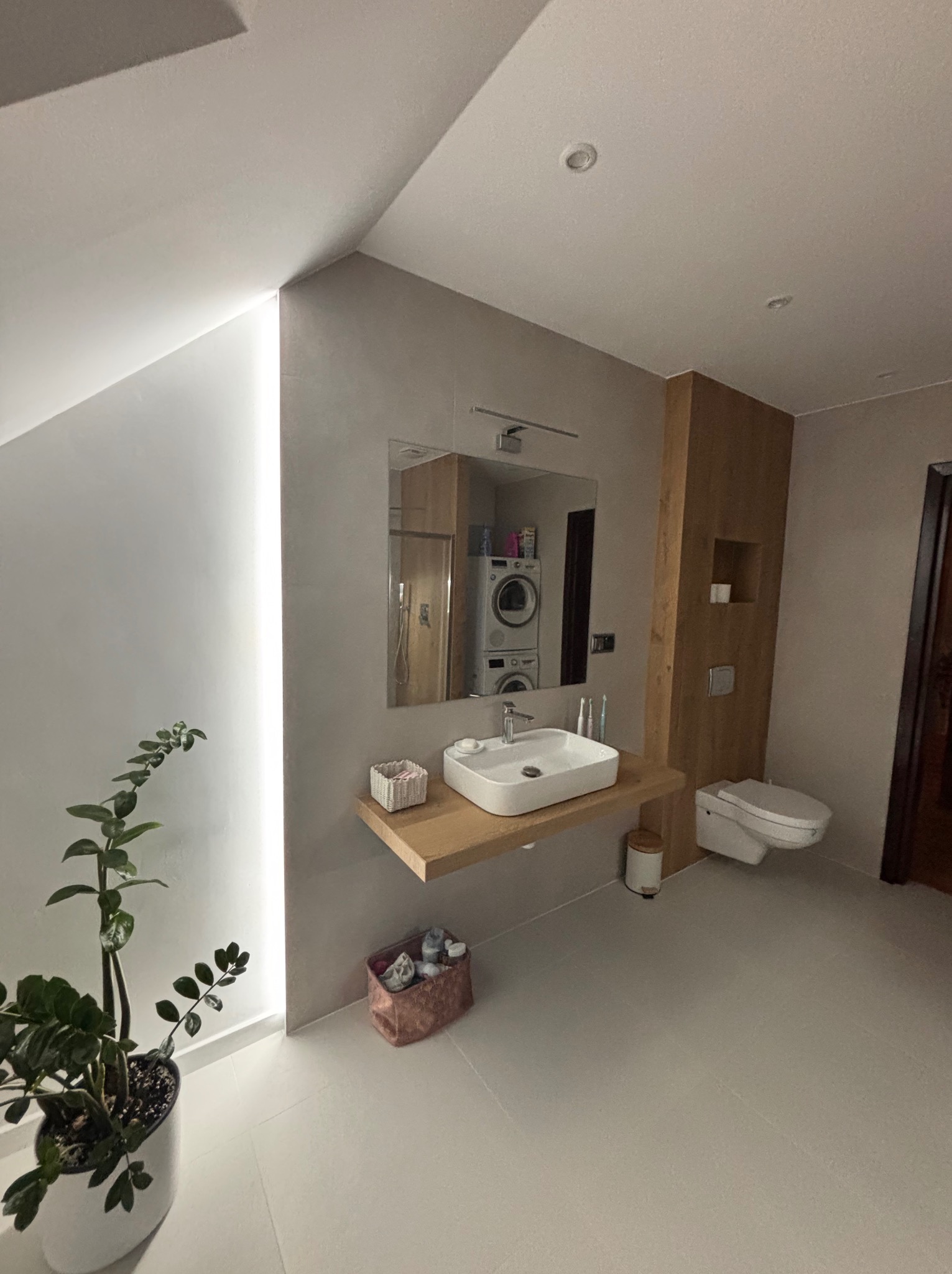 Nowoczesna łazienka z drewnianymi akcentami: umywalka na blacie, toaleta, lustro i roślina doniczkowa. W tle pralka i suszarka.