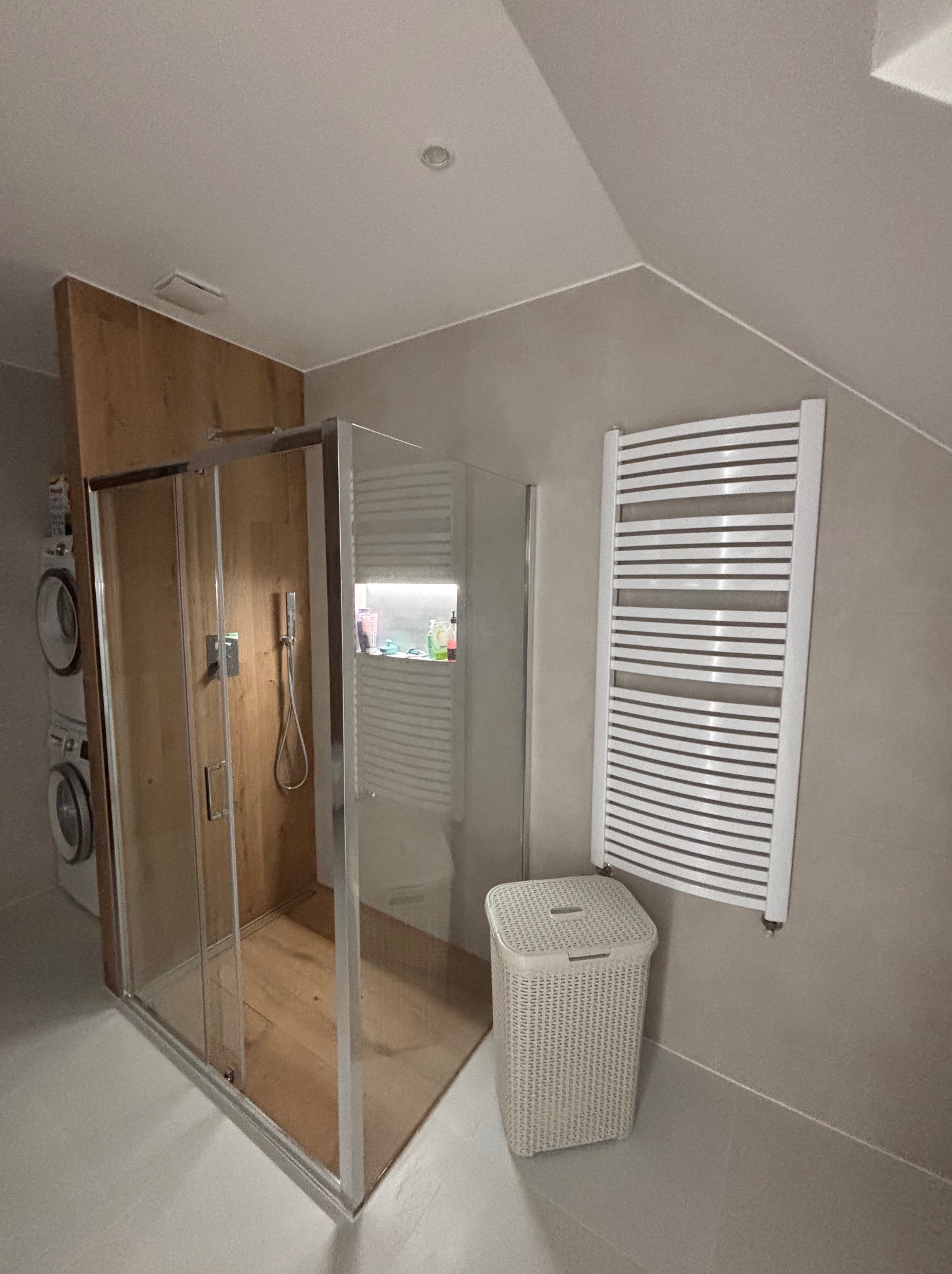 Nowoczesna łazienka na poddaszu z kabiną prysznicową typu walk-in, grzejnikiem drabinkowym i koszem na pranie. Uwagę zwraca drewniane wykończenie ściany prysznica.