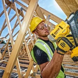 Monter w żółtym kasku i okularach ochronnych, trzymający w dłoniach akumulatorową gwoździarkę DeWalt, na tle drewnianej konstrukcji dachu i błękitnego nieba.