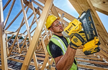 Monter w żółtym kasku i okularach ochronnych, trzymający w dłoniach akumulatorową gwoździarkę DeWalt, na tle drewnianej konstrukcji dachu i błękitnego nieba.