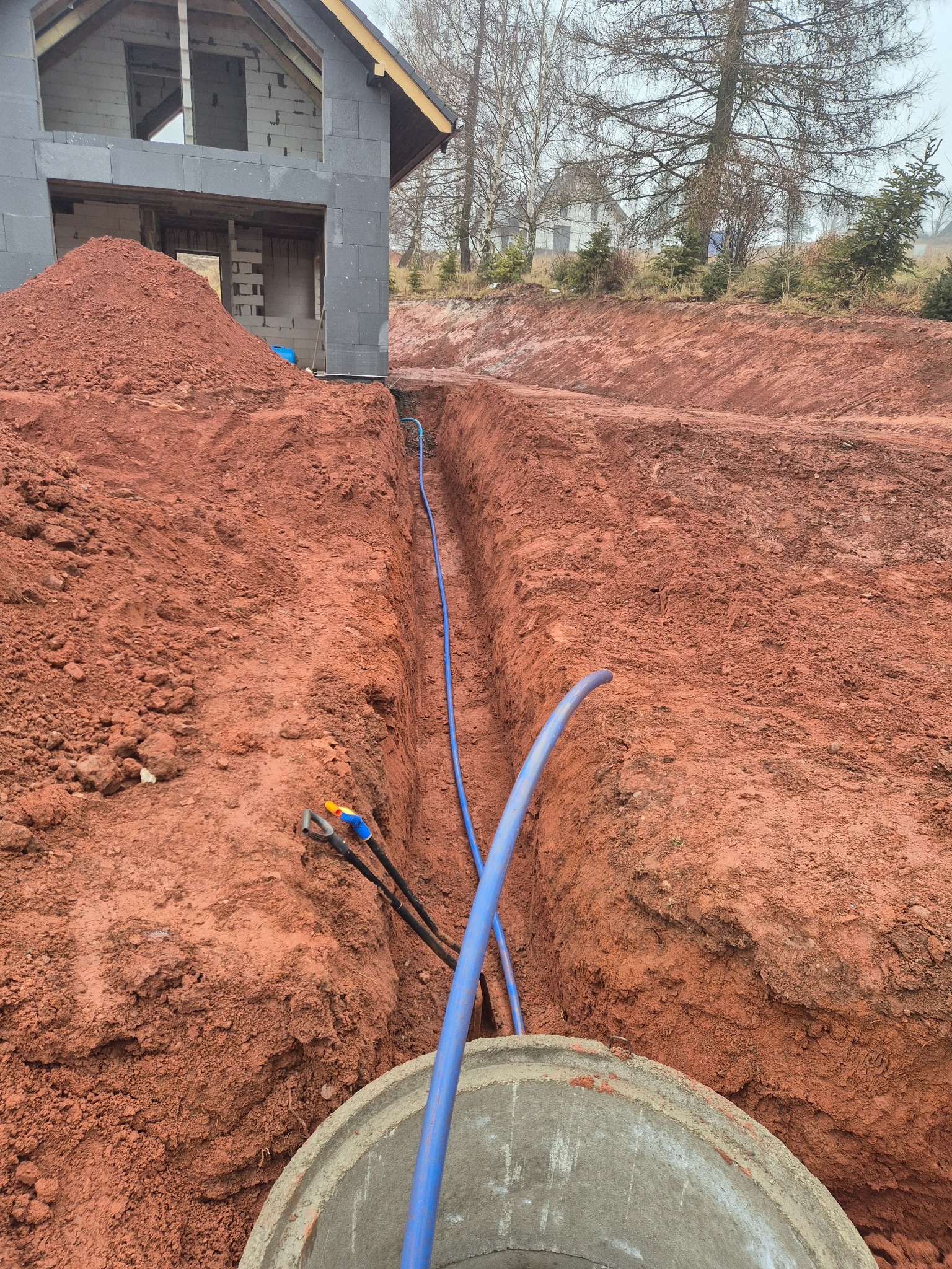 Wykop z ułożonym niebieskim rurociągiem wodnym w kierunku powstającego domu z betonu komórkowego, widoczne narzędzia i betonowy krąg studzienny.