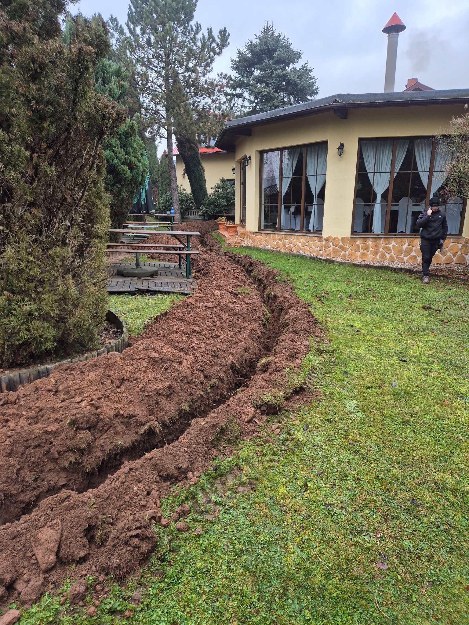 Wykopany rów w trawie na posesji, prawdopodobnie pod przyłącze wodno-kanalizacyjne, z osobą rozmawiającą przez telefon w tle.