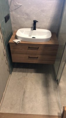 Łazienkowa szafka pod umywalkę z dwoma szufladami z drewna o jasnym odcieniu, biała umywalka nablatowa o zaokrąglonym kształcie i czarna bateria, szara płytka na podłodze, widoczna ściana z szarego...