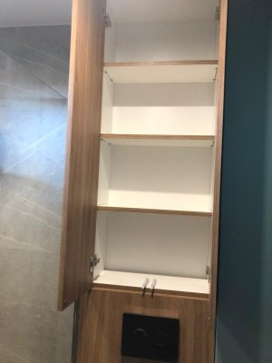 Otwarta szafka łazienkowa z drewnopodobnymi frontami i białym wnętrzem, zamontowana nad zabudową WC, z widocznymi chromowanymi uchwytami.