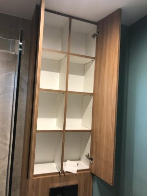 Wysoka, wąska szafka wnękowa z drewnianymi drzwiczkami i białym wnętrzem, otwarte drzwi ukazują puste półki i zawiasy, widoczny fragment łazienki z szklaną kabiną prysznicową i turkusową ścianą.