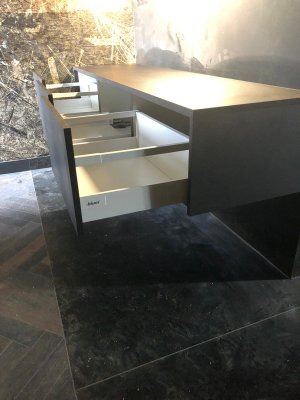 Szara szafka z wysuniętymi, białymi szufladami, widoczne okucia Blum, na podłodze z ciemnych płytek imitujących kamień, ściana w tle ze wzorem abstrakcyjnym.