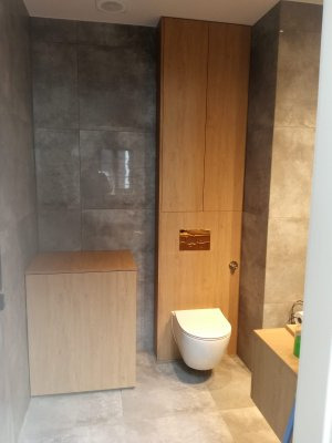 Nowoczesna łazienka z zabudową meblową z jasnego drewna: szafka pod umywalkę i wysoka szafa wnękowa nad toaletą, szara płytka na ścianach i podłodze.