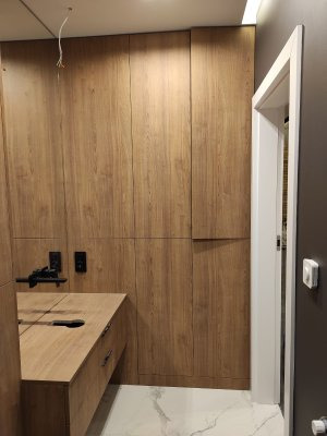 Drewniana szafa wnękowa z blatem i szufladą, widoczne czarne gniazdka elektryczne i wystające kable ze ściany, marmurowa podłoga, białe drzwi po prawej stronie.